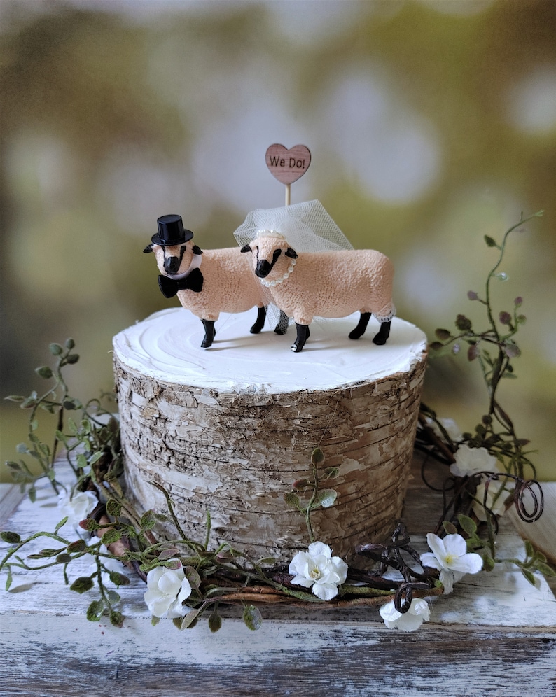 Pu&ograve; includere: Un topper per torta nuziale con due figurine di pecore, una con un cappello a cilindro e un papillon, l'altra con un velo. Il topper &egrave; posizionato su un supporto per torta in corteccia di betulla con un cartello a forma di cuore in legno che dice "We Do!"