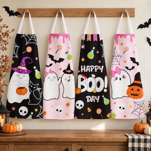 Może przedstawiać: Cztery fartuchy wiszące na drewnianym wieszaku, ozdobione wzorami halloweenowymi z duchami, dyniami, czarnymi kotami, czaszkami, pajęczynami i cukierkami. Dwa fartuchy mają czarne tło, a dwa jasnoróżowe. Jeden z nich zawiera napis HAPPY BOO DAY.