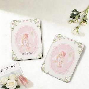 May include: Two rectangular coasters with a pink and white design. Each coaster features a central oval with a fairy illustration, surrounded by a floral border. The name "Amelia" is written on the bottom of the oval.