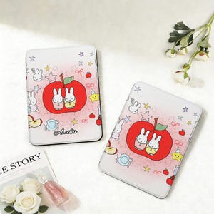 Op de afbeelding: Twee rechthoekige telefoonhoesjes met een grillig ontwerp. Elke hoes heeft een grote rode appel met twee cartoonkonijntjes, omringd door sterren, appels en andere speelse elementen. De naam "Amelia" staat op één hoesje.