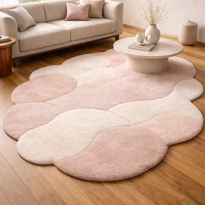 Op de afbeelding: Een groot, abstract gevormd tapijt in roze en crèmetinten. Het tapijt heeft een zacht, getextureerd oppervlak en een uniek, vloeiend ontwerp. Het ligt op een houten vloer in een woonkamer, met een bank en salontafel.