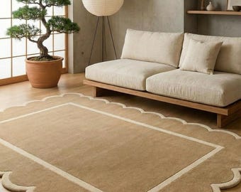May include: A neutral-toned living room featuring a beige scalloped-edge rug with a cream border. A light-colored sofa sits against a gray wall, complemented by a paper lantern floor lamp and a bonsai tree in a terracotta pot.
