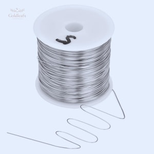 Puede incluir: Una bobina de alambre plateado con un núcleo de plástico blanco. El alambre está cuidadosamente enrollado alrededor de la bobina, con una porción del alambre extendida y con forma de patrón ondulado. La bobina tiene el número "5".