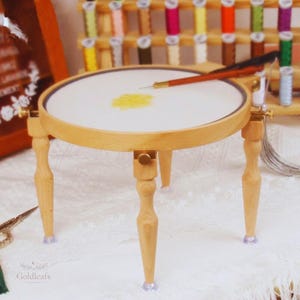 May include: A small, round wooden embroidery stand with a white fabric stretched across the hoop. A needle is inserted into the fabric, with a small patch of yellow thread visible. The stand has three legs and adjustable hardware.