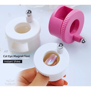 Puede incluir: Herramientas magnéticas de ojo de gato blancas y rosas para nail art. Las herramientas tienen un diseño circular con una bola plateada en la parte superior. El texto "Cat Eye Magnet Tool" y "Instant Shine" es visible. Se muestra una uña con efecto ojo de gato.