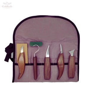 Può includere: Un set di utensili per intaglio del legno con manici in legno e lame in acciaio inossidabile, presentato in un astuccio arrotolabile in tela viola. Il set include varie sgorbie, coltelli e un utensile a gancio.