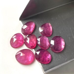 May include: A collection of seven faceted, oval-shaped pinkish-red gemstones with a polished finish. The stones are arranged in a cluster on a metallic silver-coloured scoop, displaying translucent qualities and geometric light reflections on their surfaces.
