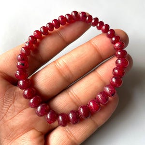 Puede incluir: Una pulsera elástica compuesta por numerosas cuentas pequeñas y redondas de piedra preciosa de color rojo intenso. Cada cuenta presenta una superficie pulida y ligeramente translúcida con variaciones sutiles de color e inclusiones oscuras.
