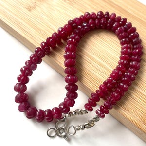 Può includere: Una collana composta da numerose perline circolari scanalate di colore rosso intenso infilate insieme. Il filo presenta un meccanismo di chiusura color argento a un'estremità, costituito da un moschettone e diverse piccole perline argentate lisce.