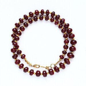 Puede incluir: Un collar que presenta una serie de cuentas circulares pulidas de color rojo intenso que se alternan con pequeños espaciadores esféricos de metal dorado. El hilo está dispuesto en un bucle circular e incluye un cierre de mosquetón dorado.