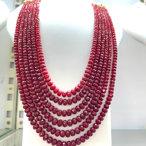 Puede incluir: Un collar de múltiples hilos con siete capas de cuentas de piedras preciosas de color rojo oscuro facetadas. Los hilos están dispuestos en un diseño en cascada graduado, asegurados en la parte superior con cierres de metal dorado. Cada cuenta es pulida.