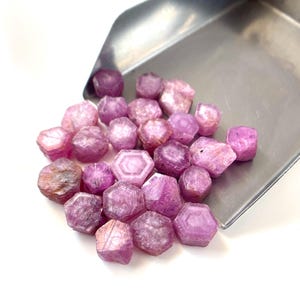 May include: A collection of small, raw, hexagonal-shaped pink and magenta gemstones poured from a stainless steel metal scoop. The stones exhibit varying levels of translucency, natural crystalline textures, and distinct geometric facets.