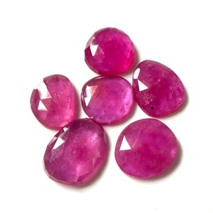 Puede incluir: Un conjunto de cinco rubíes sueltos, facetados y de color rosa vibrante con un acabado pulido. Cada piedra preciosa presenta una forma redondeada e irregular con múltiples facetas planas que reflejan la luz, mostrando un color rosa magenta profundo y saturado.