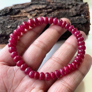 Puede incluir: Una pulsera elástica compuesta por numerosas cuentas pequeñas y redondas de piedra preciosa de color rojo intenso. Las cuentas presentan un acabado liso y pulido con sutiles variaciones en la intensidad del color, formando un círculo uniforme.