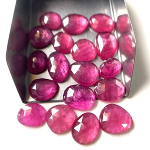 May include: A collection of faceted, oval-shaped magenta gemstones with a polished finish. Several stones are contained within a small, silver-colored metal scoop, while others are scattered below it on a plain white surface.