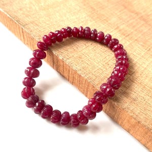 May include: A circular bracelet composed of numerous small, deep red, carved gemstone beads. Each bead features a distinct ribbed or pumpkin-like texture, strung together to form a continuous loop of uniform, dark crimson, faceted-style stones.