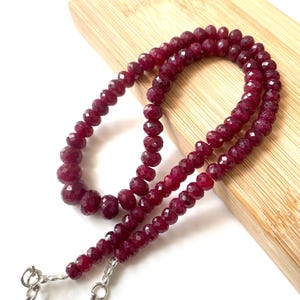 May include: A necklace composed of faceted, deep red, circular beads arranged in a single strand. The beads exhibit a consistent dark crimson hue and a reflective, multi-faceted surface. A silver-toned metal clasp is attached to the end of the strand.