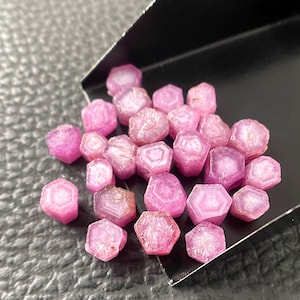 May include: A collection of small, hexagonal pink corundum crystals with visible internal geometric banding patterns. The stones are clustered together on a black metal scoop, displaying translucent to opaque surfaces with varying shades of magenta.