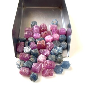 Pode incluir: Uma pá de metal cheia com uma coleção de pequenas pedras preciosas brutas e não lapidadas em tons de rosa profundo, magenta e azul-acinzentado suave. As pedras têm texturas irregulares, facetadas e cristalinas como minerais soltos.