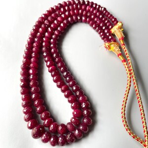 May include: A multi-strand necklace featuring deep red, faceted, round beads. The strands are gathered at the ends with gold-colored metallic thread wrappings, which connect to a braided red and gold cord for securing the necklace around the neck.