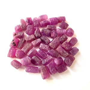 Puede incluir: Una colección de pequeñas piedras preciosas de rubí en bruto, de forma cilíndrica y con un vibrante tono rosa magenta. Cada piedra presenta una superficie rugosa y texturizada con variaciones naturales en forma y opacidad, agrupadas.