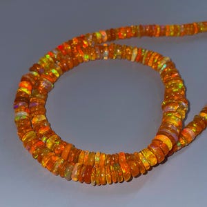 May include: A strand of small, circular, orange-toned opal beads. Each bead features a translucent quality with internal flashes of yellow, green, and red light. The beads are strung together in a continuous line, forming a curved, circular shape.
