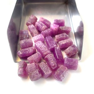 May include: A collection of small, raw, unpolished ruby gemstones with a vibrant magenta-pink hue. These natural, rough-cut stones are scattered inside a metallic silver-colored scoop, displaying varied crystalline textures and opaque surfaces.