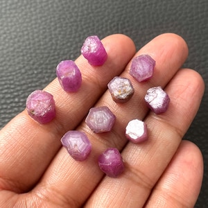 May include: A collection of ten small, raw, hexagonal-shaped pinkish-red rubies. Each gemstone displays a natural, unpolished texture with visible crystalline structures and varying degrees of opacity, showcasing a distinct six-sided geometric form.