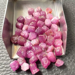 May include: A collection of small, raw, unpolished pink and magenta gemstones with irregular, faceted shapes. These loose stones are contained within a metallic, silver-colored scoop, displaying varied textures and translucent to opaque surfaces.