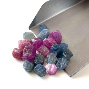 May include: A collection of small, raw, uncut gemstones in shades of deep blue and vibrant pink, resting on a metallic silver scoop. The stones have irregular, faceted surfaces and varying levels of translucency, displaying natural textures.