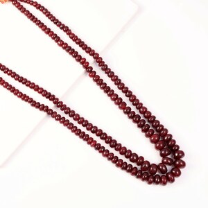 May include: A long, double-strand necklace composed of small, smooth, round, deep red beads. The beads are uniform in size and shape, strung together to create a continuous, flexible chain that loops to form two distinct, parallel layers.