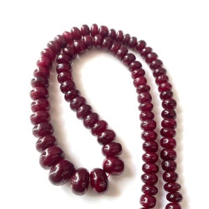May include: A strand of deep red, polished, disc-shaped beads arranged in a curved line. The beads have a smooth, glossy texture and are strung together on a thin red cord, creating a continuous loop of uniform, rounded, dark crimson gemstone pieces.