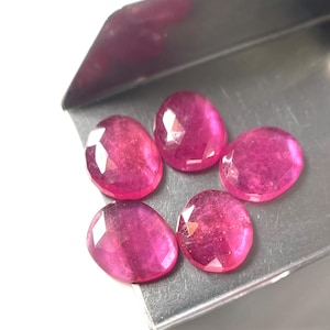 Puede incluir: Cinco piedras preciosas rosas de forma ovalada y facetadas, dispuestas en un grupo sobre una pala metálica. Cada piedra muestra un tono magenta translúcido y vibrante con múltiples facetas geométricas que reflejan la luz.
