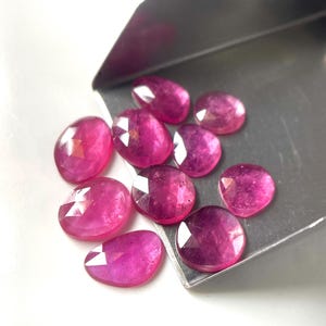 Puede incluir: Una colección de diez rubíes rosas facetados de forma ovalada que descansan sobre una pala de metal plateado. Las piedras preciosas exhiben un tono magenta vibrante y translúcido con inclusiones internas visibles y superficies pulidas.