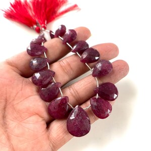 Peut inclure: Un collier de pierres précieuses rubis en forme de poire, facettées et d'un magenta profond, disposées en goutte. Les pierres sont enfilées sur un fil fin et terminées par un pompon décoratif rouge vif, offrant une surface polie et brillante.