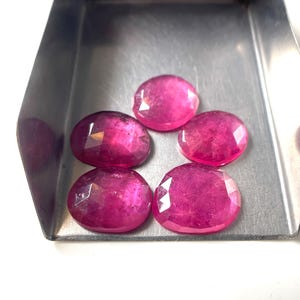 May include: Five oval-shaped, faceted pink rubies arranged in a cluster inside a metallic silver-coloured scoop. Each gemstone displays a translucent quality with visible internal inclusions and a polished, multi-faceted surface reflecting light.