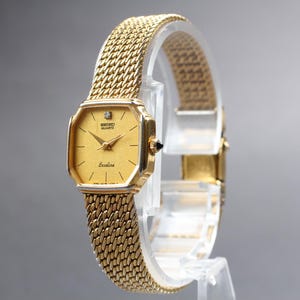 Puede incluir: Reloj Seiko Quartz Exceline dorado con una correa de malla. El reloj tiene una esfera cuadrada dorada con un pequeño diamante y agujas doradas. El reloj se exhibe sobre un soporte transparente.