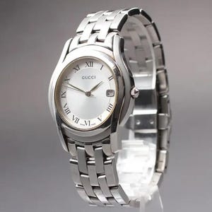 May include: A silver-toned Gucci wristwatch with a round face and a linked metal band. The watch face has Roman numeral hour markers and the Gucci logo. The date is displayed at the 3 o'clock position.