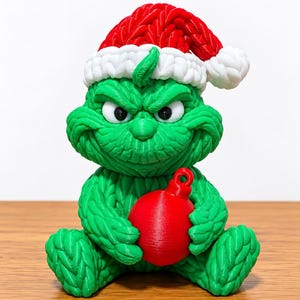 May include: A green, textured figurine of a creature with a scowling expression, wearing a red and white Santa hat. The figure has a knitted-style surface texture and is holding a smooth, round, red Christmas ornament in its hands.