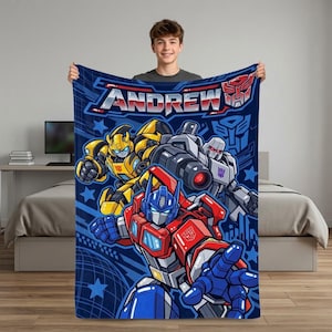 Könnte beinhalten: Eine rechteckige Fleecedecke mit dunkelblauem Hintergrund und dem Namen ANDREW in großen silbernen Buchstaben. Das Design zeigt farbenfrohe Illustrationen der Transformers-Charaktere Bumblebee, Megatron und Optimus Prime.