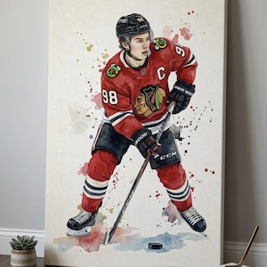 May include: Watercolor painting of a hockey player in a red and black Chicago Blackhawks jersey with the number 98. The player is holding a hockey stick and a puck is on the ice. The artwork has colorful paint splatter accents.