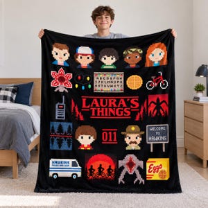 May include: A black fleece blanket featuring a pixelated graphic print of characters and icons from the show Stranger Things, including the text LAURA'S THINGS, an alphabet wall with lights, a bicycle, a van, and a box of Eggo waffles.