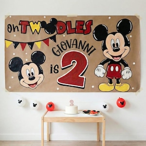 Puede incluir: Una pancarta marrón con las palabras "oh TWODDLES" y "GIOVANNI is 2" en rojo y negro. Mickey Mouse aparece en la pancarta y en los globos. Una pequeña tarta blanca con una vela con el número 2 está sobre una mesa.