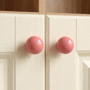 Puede incluir: Dos pomos de armario esf&eacute;ricos y lisos de color rosa empolvado est&aacute;n montados en puertas de armario de color crema claro. Las puertas presentan una sutil textura de veta de madera y un dise&ntilde;o de panel elevado, ofreciendo una instalaci&oacute;n limpia.