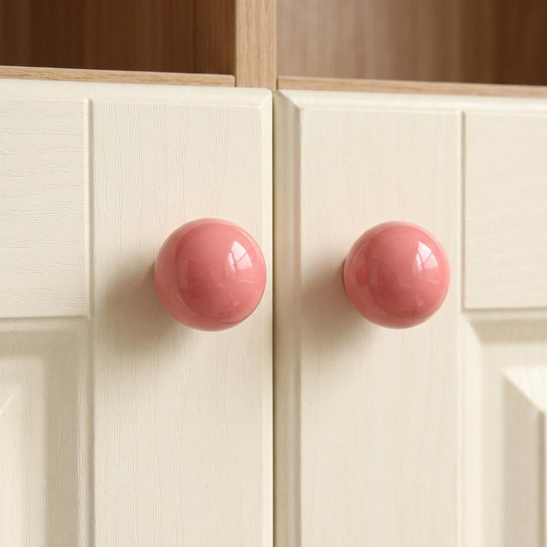 Puede incluir: Dos pomos de armario esf&eacute;ricos y lisos de color rosa empolvado est&aacute;n montados en puertas de armario de color crema claro. Las puertas presentan una sutil textura de veta de madera y un dise&ntilde;o de panel elevado, ofreciendo una instalaci&oacute;n limpia.