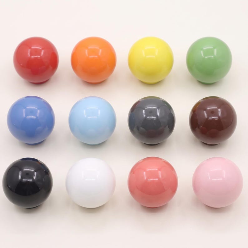 Puede incluir: Una colecci&oacute;n de doce pomos esf&eacute;ricos para armarios dispuestos en una cuadr&iacute;cula. Los pomos presentan un acabado liso y brillante y vienen en una variedad de colores s&oacute;lidos que incluyen rojo, naranja, amarillo, verde, azul, azul claro, gris, marr&oacute;n, negro, blanco, coral y rosa.