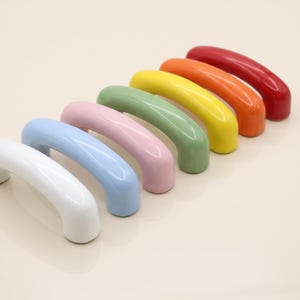 May include: A set of seven arched cabinet handles arranged in a diagonal row, featuring a smooth, glossy finish in a spectrum of colours including white, light blue, soft pink, sage green, yellow, orange, and deep red, each with two mounting points.