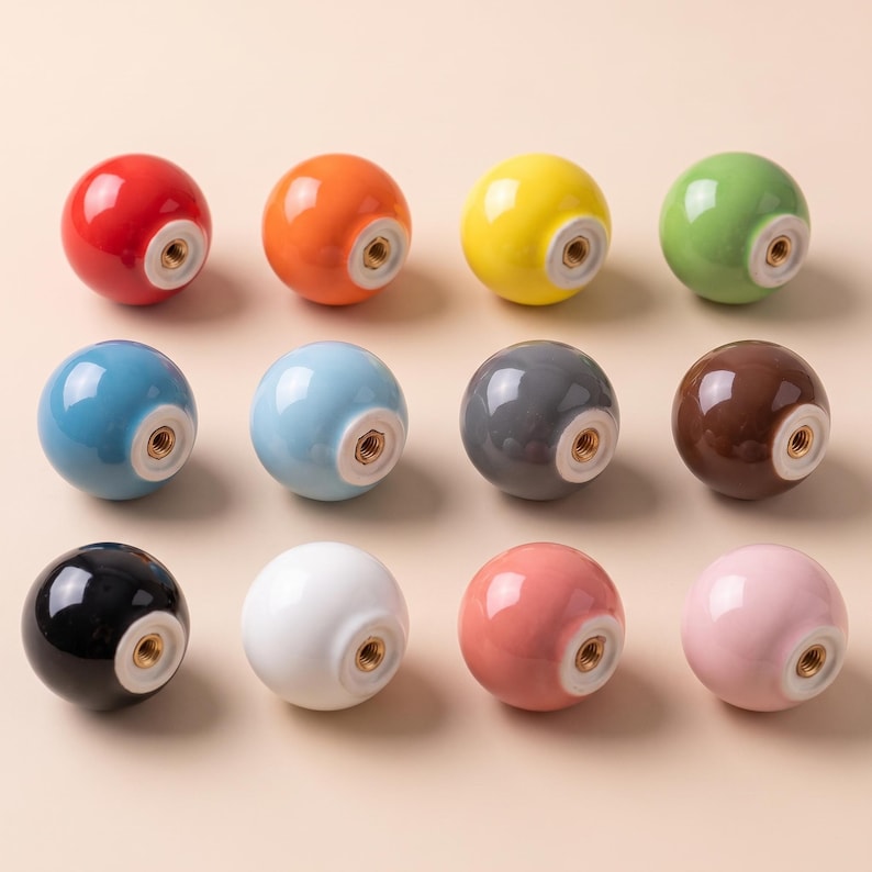 Puede incluir: Una colecci&oacute;n de doce pomos esf&eacute;ricos para armarios en varios colores s&oacute;lidos, incluyendo rojo, naranja, amarillo, verde, azul, azul claro, gris, marr&oacute;n, negro, blanco, coral y rosa. Cada pomo tiene un acabado brillante y un orificio central.
