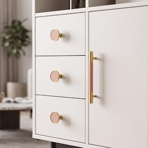 May include: A white cabinet featuring three drawers with round, gold-rimmed pink knobs and a single vertical door handle with a gold frame and a pink center insert. The cabinet surface is smooth and matte, showcasing a clean, functional design.