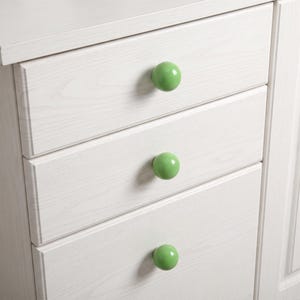 Puede incluir: Una c&oacute;moda de madera blanca con tres cajones apilados, cada uno equipado con un pomo redondo, liso y de color verde claro. La veta de la madera es visible en los frentes de los cajones, aportando una apariencia texturizada al mueble.